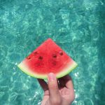 Hand holding a ripe watermelon slice with a turquoise pool background, perfect summer vibes.